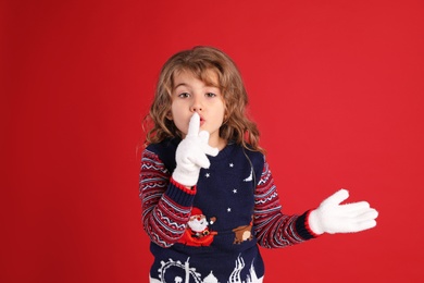 Photo of Cute little girl in Christmas sweater showing silence gesture against red background