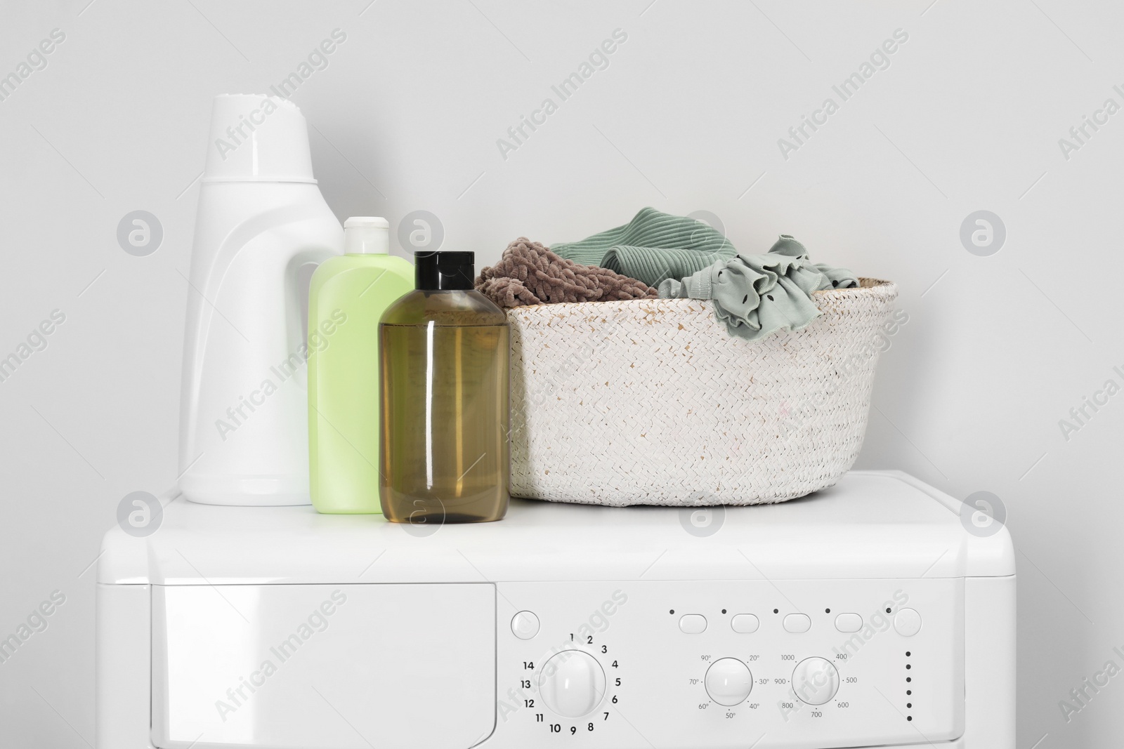 Photo of Baby clothes in wicker basket and laundry detergents on washing machine near light wall