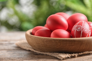 Photo of Wooden bowl with red painted Easter eggs on table against blurred background, space for text