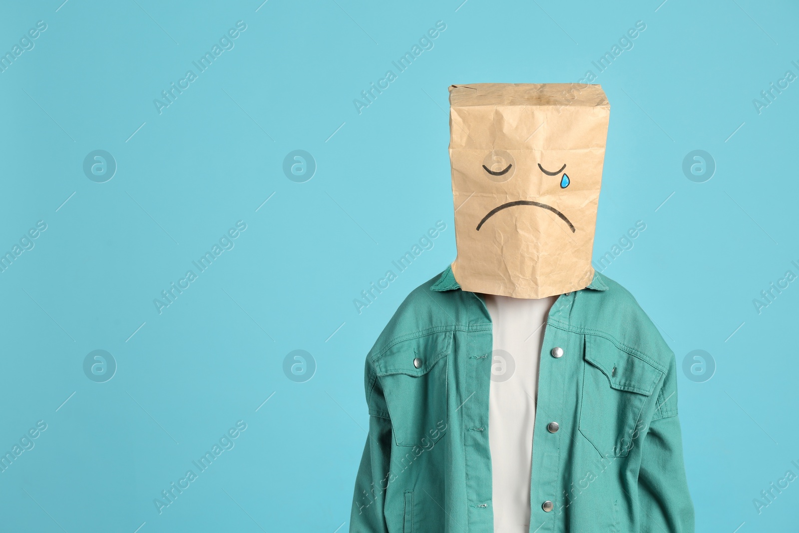 Photo of Woman wearing paper bag with drawn sad face on light blue background. Space for text