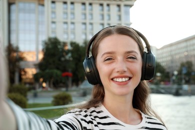 Photo of Portrait of smiling woman in headphones taking selfie on city street