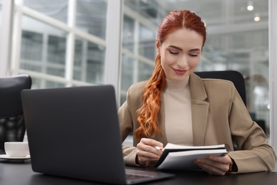 Happy woman with notebook working near laptop at black desk in office