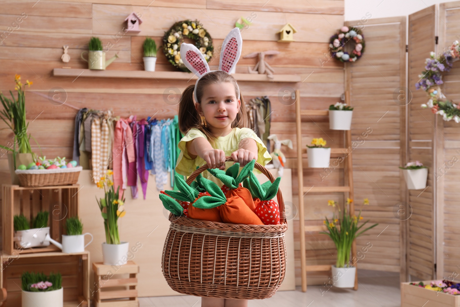 Photo of Adorable little girl with bunny ears and basket full of toy carrots in Easter photo zone