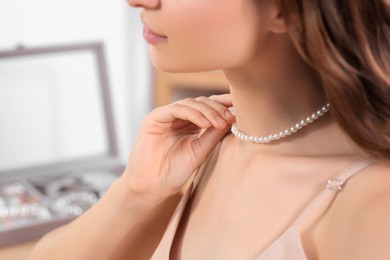 Young woman trying on elegant pearl necklace indoors, closeup