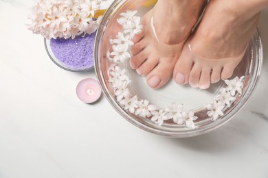 Photo of Woman soaking her feet in bowl with water and flowers on white marble table, top view. Pedicure procedure