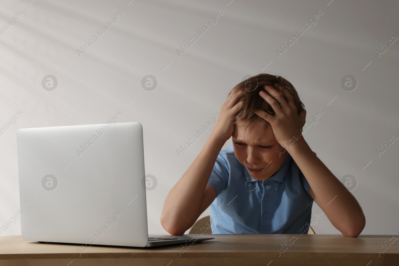 Photo of Upset boy with laptop at wooden table. Cyber bullying