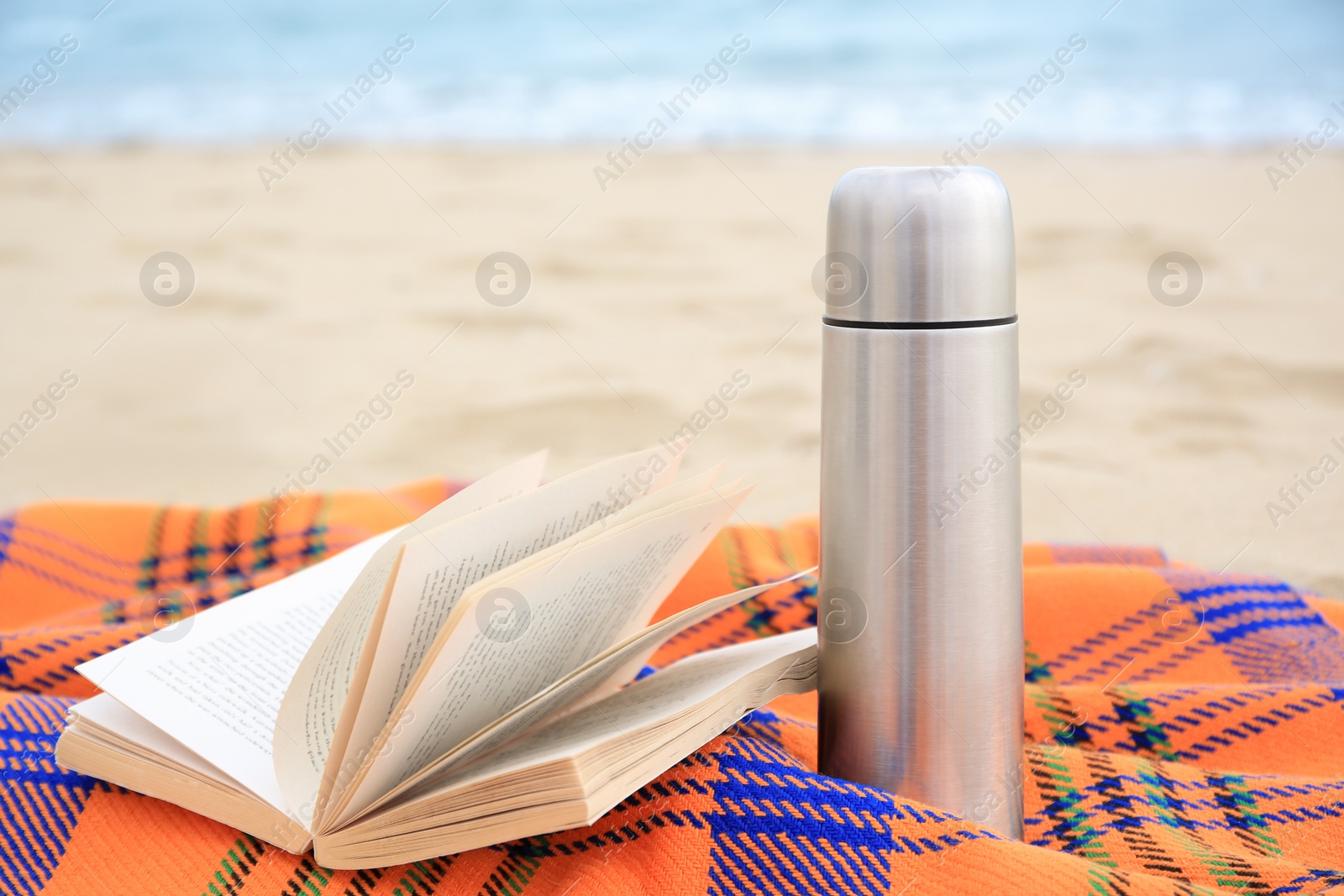 Photo of Metallic thermos with hot drink, open book and plaid on sandy beach near sea