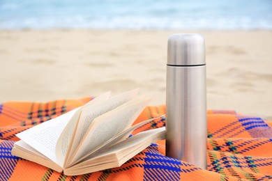 Metallic thermos with hot drink, open book and plaid on sandy beach near sea