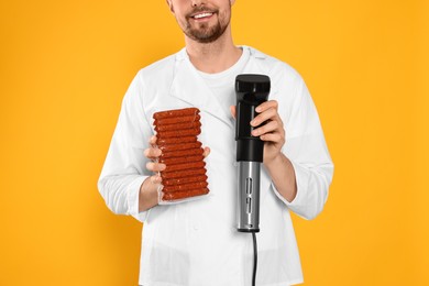 Smiling chef holding sous vide cooker and sausages in vacuum pack on orange background, closeup