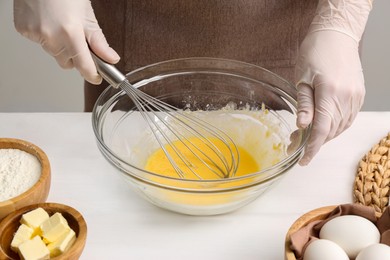 Woman whisking crepe batter in glass bowl at white wooden table, closeup
