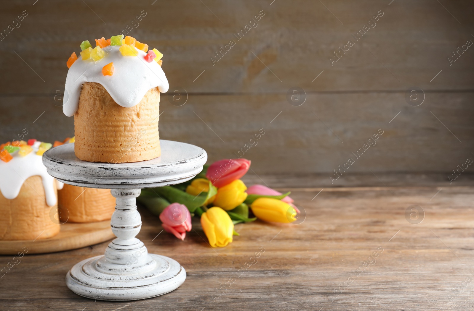 Photo of Stand with traditional Easter cake and beautiful tulips on wooden table. Space for text