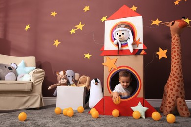 Photo of Cute little boy playing with cardboard rocket and toys at home. Child's room interior