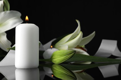 White lilies and burning candles on black mirror surface in darkness, closeup with space for text. Funeral symbols