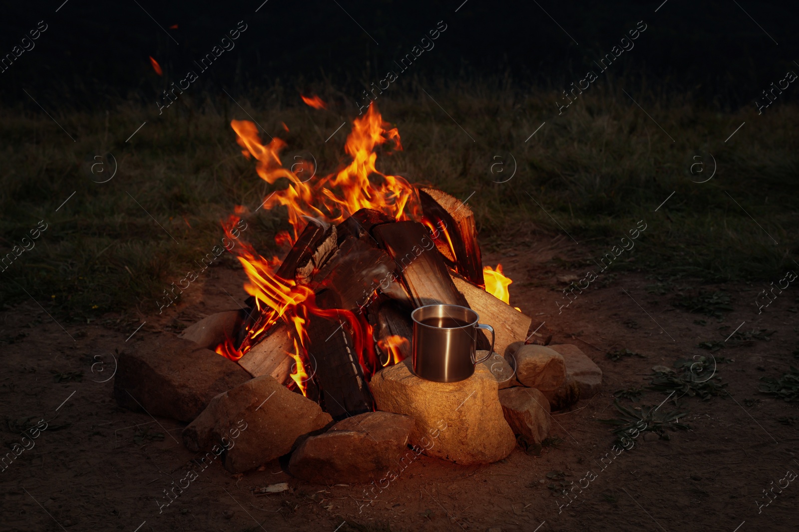 Photo of Metal mug with drink near beautiful bonfire outdoors at night. Camping season
