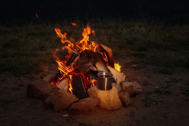 Photo of Metal mug with drink near beautiful bonfire outdoors at night. Camping season