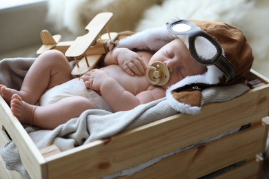 Cute newborn baby wearing aviator hat with toy sleeping in wooden crate