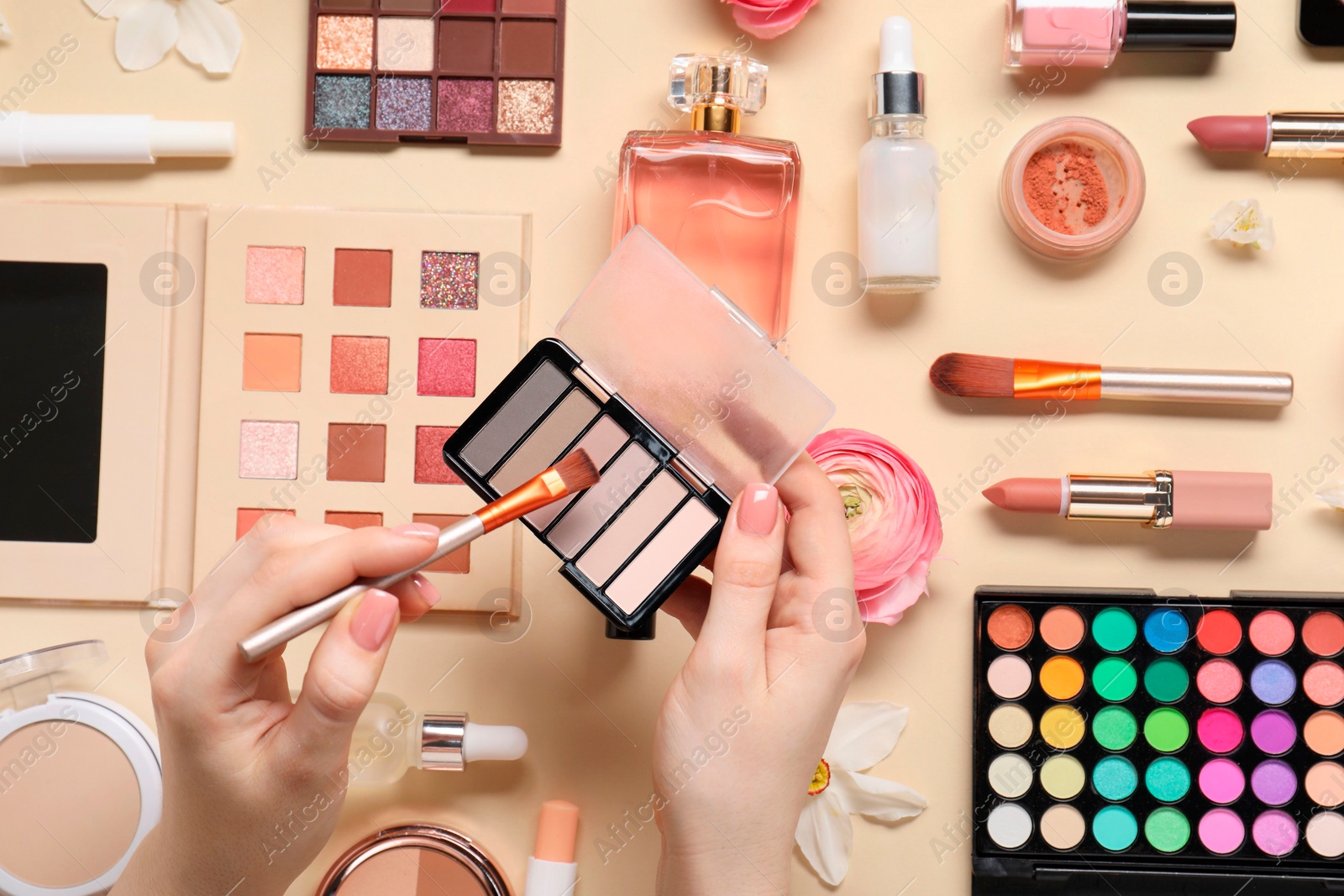 Photo of Woman applying eye shadow palette on brush, closeup. Different makeup products and beautiful spring flowers on beige background, top view