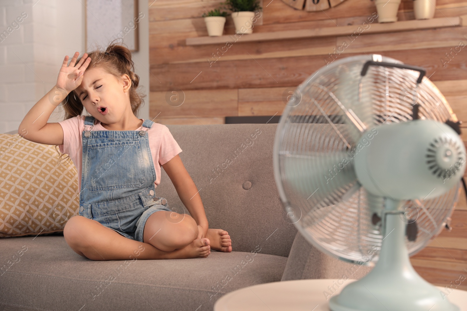 Photo of Little girl enjoying air flow from fan on sofa in living room. Summer heat