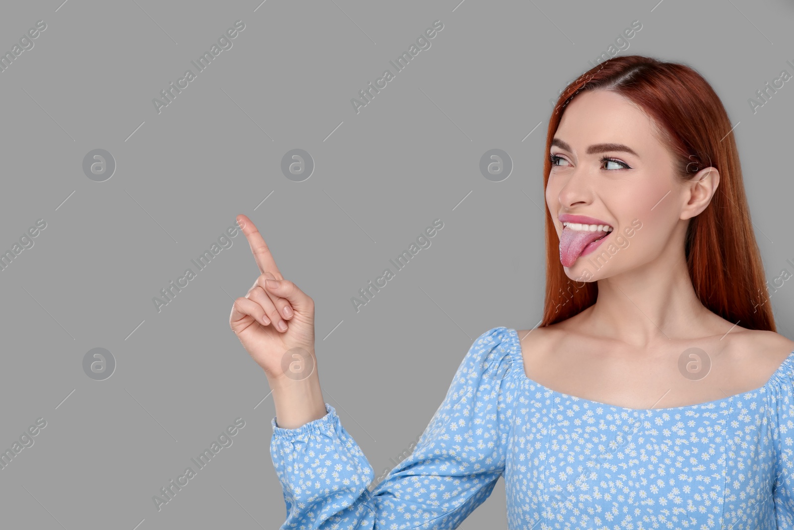Photo of Happy woman showing her tongue and pointing at something on gray background. Space for text