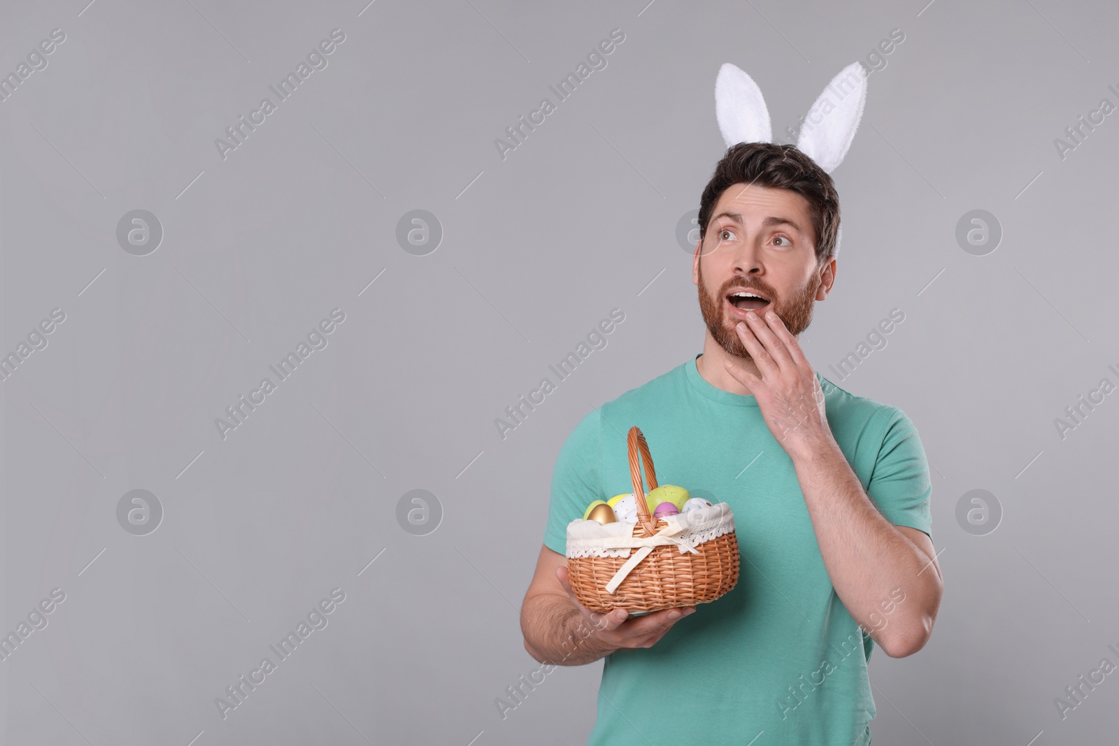 Photo of Emotional man in cute bunny ears headband holding wicker basket with Easter eggs on light grey background. Space for text