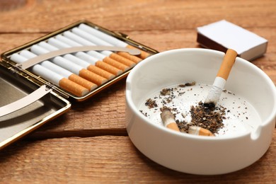 Cigarettes in case and ashtray with stubs on wooden table, closeup