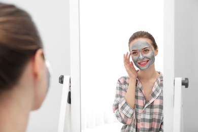 Young woman with cleansing mask on her face near mirror in bathroom. Skin care