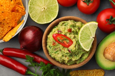 Photo of Bowl of delicious guacamole, nachos chips and ingredients on grey table, flat lay