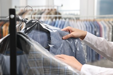 Photo of Dry-cleaning service. Woman taking jacket in plastic bag from rack indoors, closeup
