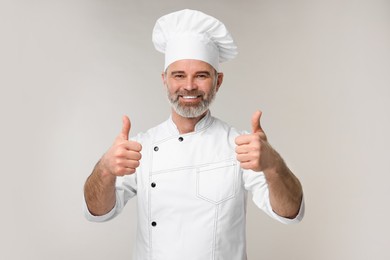 Happy chef in uniform showing thumbs up on grey background