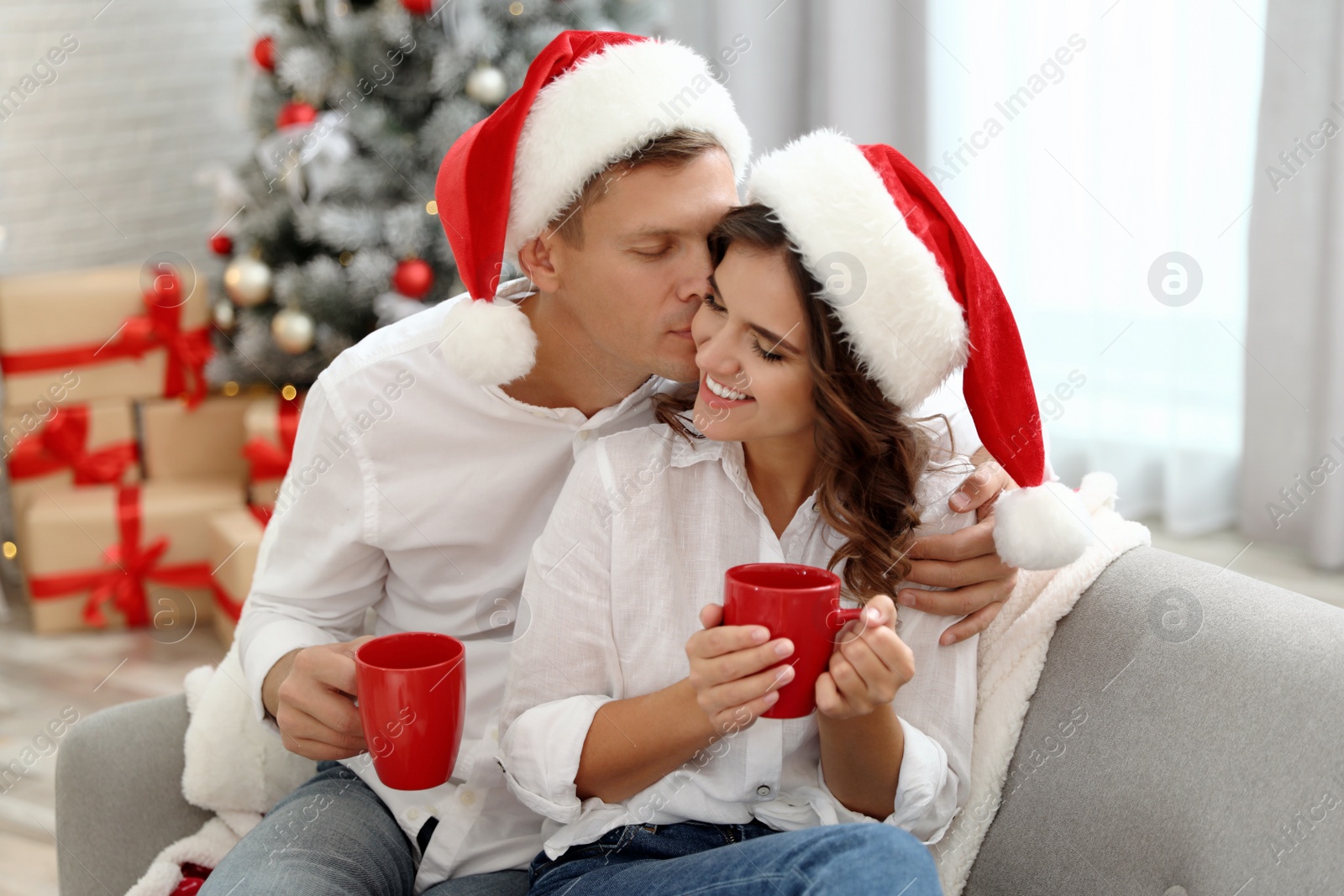 Image of Happy couple in Santa hats with cups of hot drink near Christmas tree at home