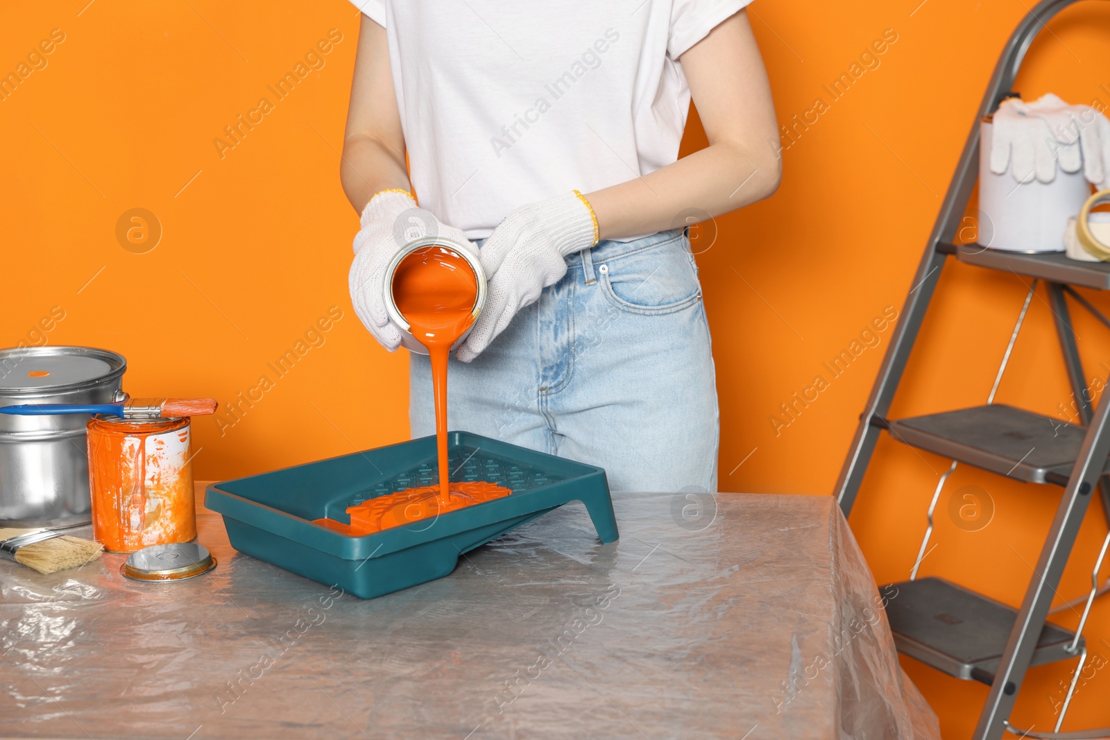 Photo of Woman pouring orange paint from can into tray at table, closeup