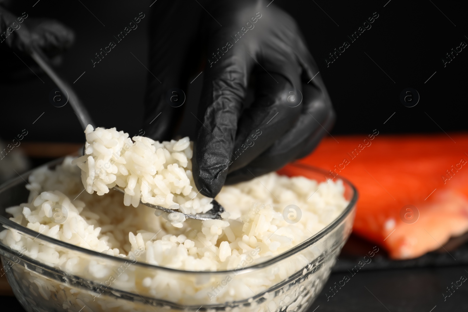 Photo of Chef in gloves taking cooked rice for sushi with spoon at dark table, closeup