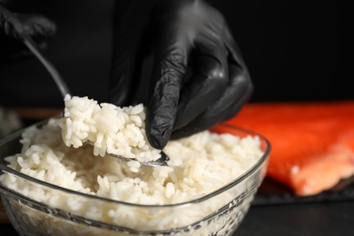 Photo of Chef in gloves taking cooked rice for sushi with spoon at dark table, closeup