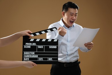 Photo of Emotional asian actor performing role while second assistant camera holding clapperboard on brown background, selective focus. Film industry