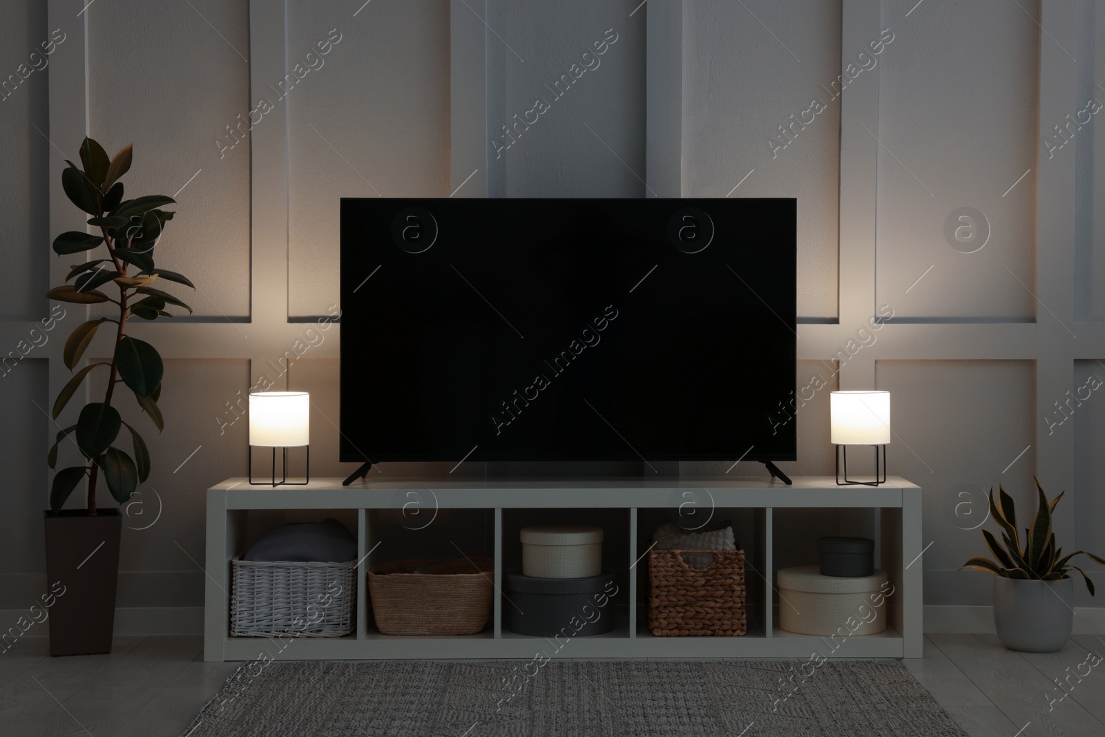 Photo of Modern TV on cabinet, lamps and beautiful houseplants near white wall in room. Interior design