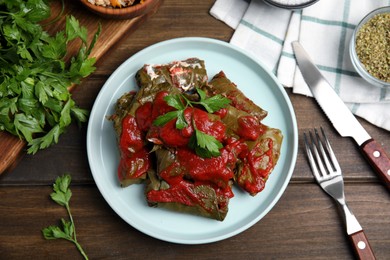 Photo of Plate of delicious stuffed grape leaves with tomato sauce, parsley and cutlery on wooden table, flat lay