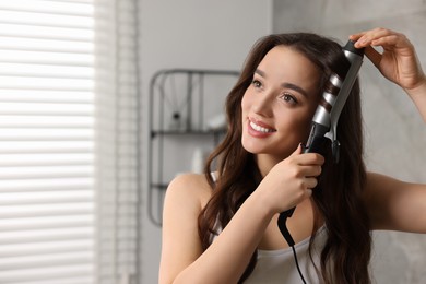 Smiling woman using curling hair iron in bathroom. Space for text