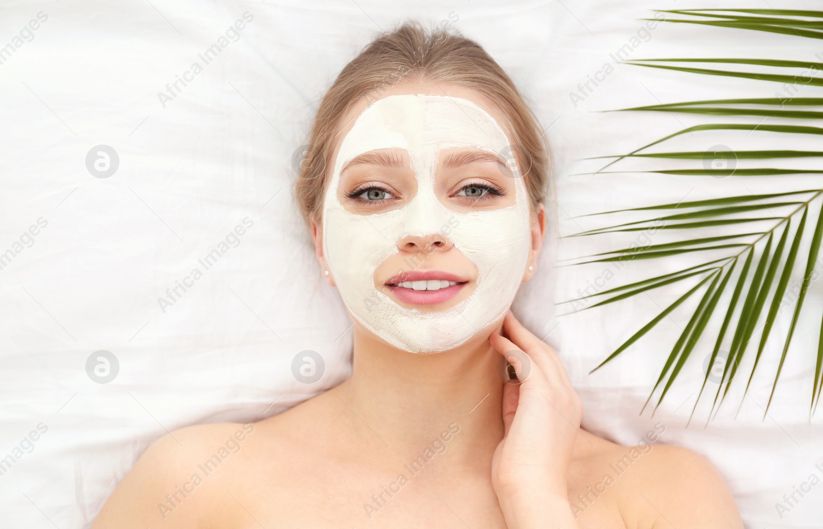 Photo of Beautiful woman with clay facial mask and tropical leaf on white fabric, above view