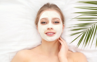 Beautiful woman with clay facial mask and tropical leaf on white fabric, above view