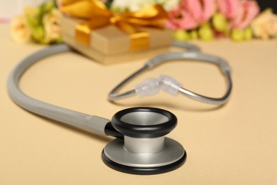 Photo of Stethoscope, gift box and eustoma flowers on beige background, closeup. Happy Doctor's Day