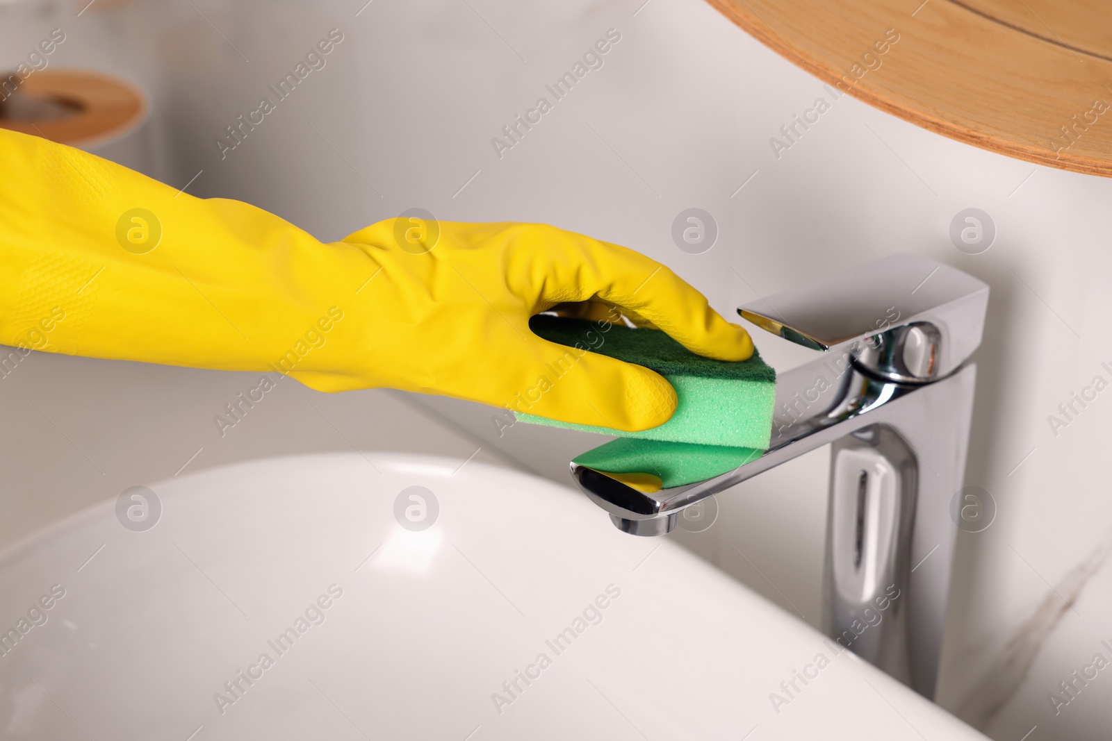 Photo of Woman in gloves cleaning faucet of bathroom sink with sponge, closeup