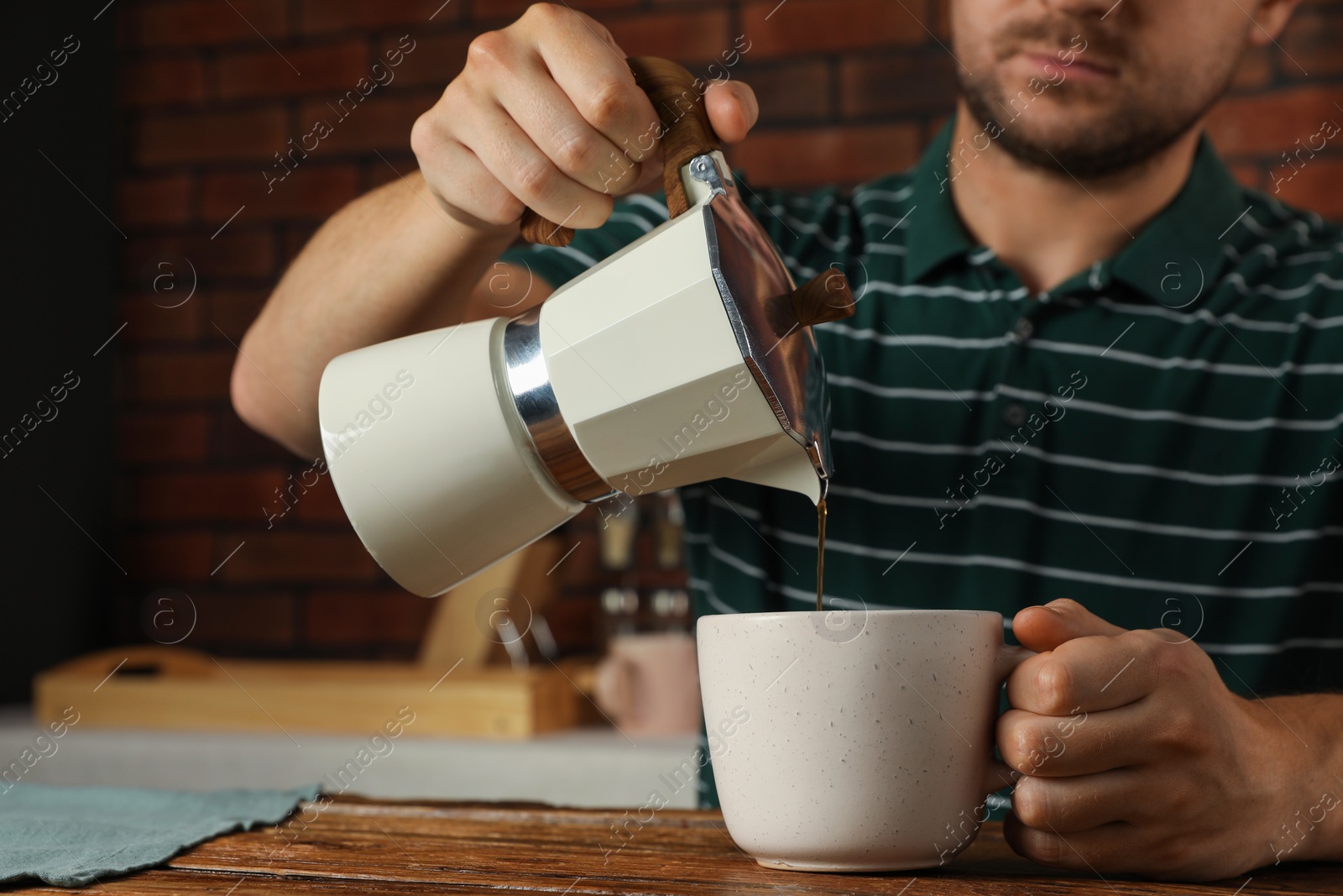 Photo of Man pouring aromatic coffee from moka pot into cup at wooden table indoors, closeup