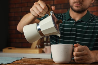 Man pouring aromatic coffee from moka pot into cup at wooden table indoors, closeup