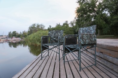 Camouflage fishing chairs on wooden pier near river