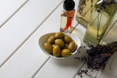 Photo of Bottles of different cooking oils, lavender flowers and olives on white wooden table