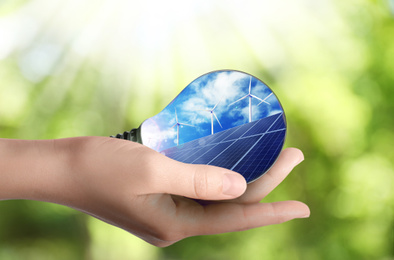 Alternative energy source. Woman holding light bulb with solar panels and wind turbines, closeup