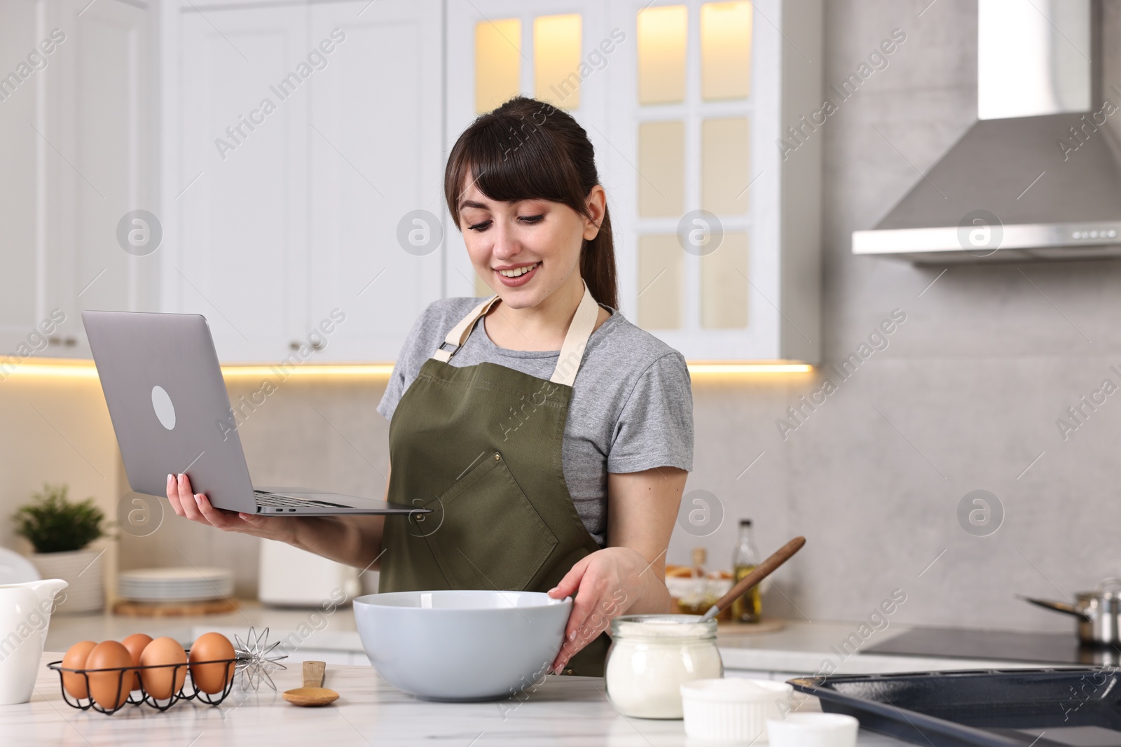 Photo of Happy young housewife with laptop cooking at white marble table in kitchen