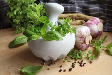 Mortar with different fresh herbs near garlic, horseradish roots and black peppercorns on wooden table, closeup