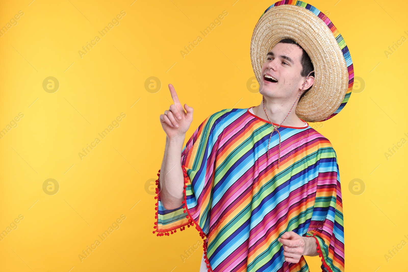 Photo of Young man in Mexican sombrero hat and poncho pointing at something on yellow background. Space for text