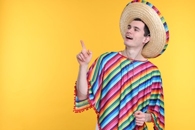 Young man in Mexican sombrero hat and poncho pointing at something on yellow background. Space for text